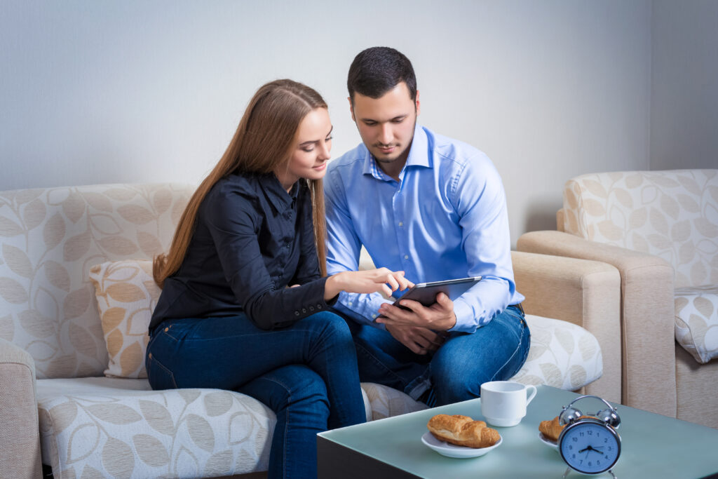 A young couple sits on a sofa, sharing a tablet while enjoying coffee and pastries.