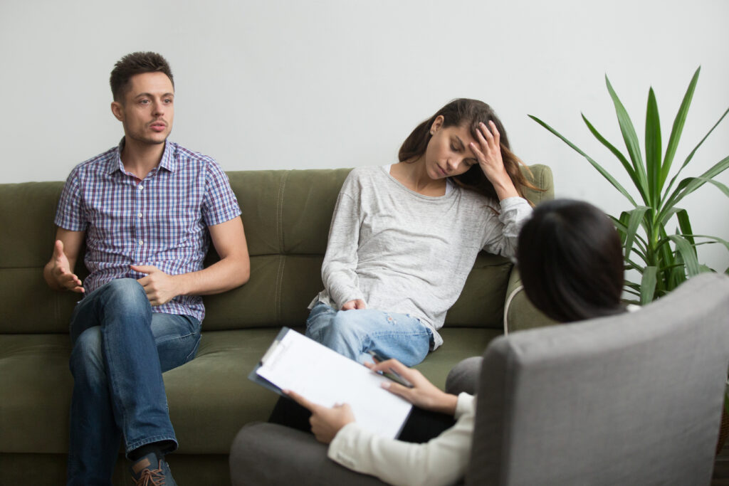 A couple attends a therapy session, with the man speaking while the woman appears stressed and holds her forehead as a therapist listens and takes notes.