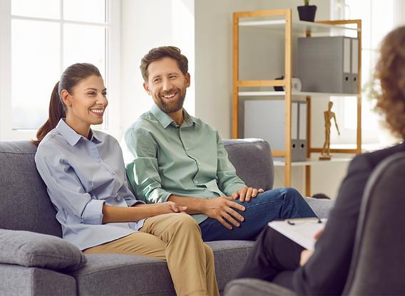 A smiling couple sits on a sofa while speaking with a therapist.
